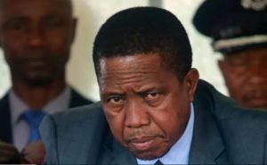 A solemn crowd gathers near a Zambian flag at half-mast, mourning former President Edgar Lungu.
