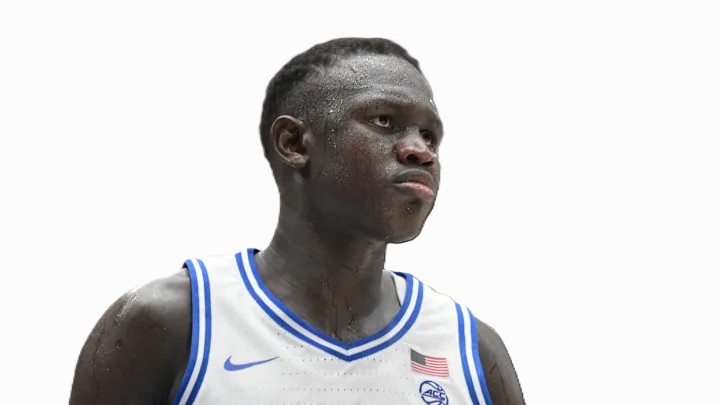 Khaman Maluach on stage at the NBA Draft, smiling in a suit lined with South Sudan and Uganda flags, holding his Phoenix Suns jersey beside Commissioner Adam Silver.