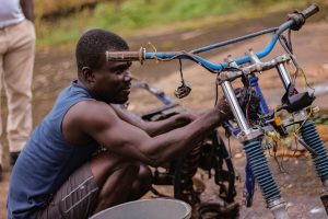 Ugandan health worker riding a Buffalo Bicycle along a dirt road, carrying medical supplies through a rural village.
