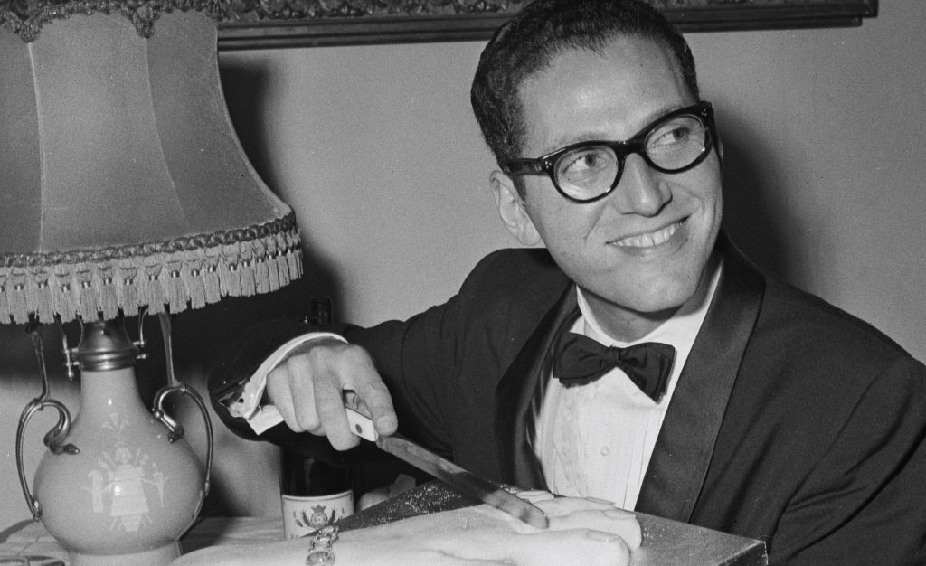 Portrait of Tom Lehrer smiling beside a piano, representing his legacy in music and satire.