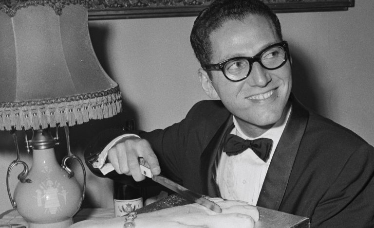 Portrait of Tom Lehrer smiling beside a piano, representing his legacy in music and satire.