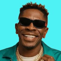 Shatta Wale performing on stage with a Ghanaian flag draped over his shoulders as fans wave and cheer in a crowded stadium.