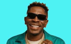 Shatta Wale performing on stage with a Ghanaian flag draped over his shoulders as fans wave and cheer in a crowded stadium.