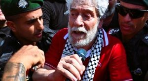 A bearded Georges Abdallah, holding papers, exits French prison after 41 years, greeted by supporters.