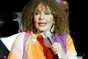 Dame Cleo Laine performing on stage in her later years, smiling with a microphone in hand. Ask ChatGPT