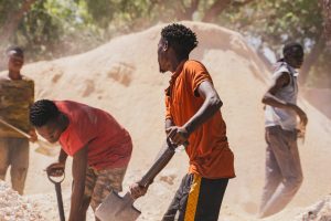A group of people digging with basic tools and hands in a muddy, chaotic mining site surrounded by makeshift tents and wooden structures. Ask ChatGPT